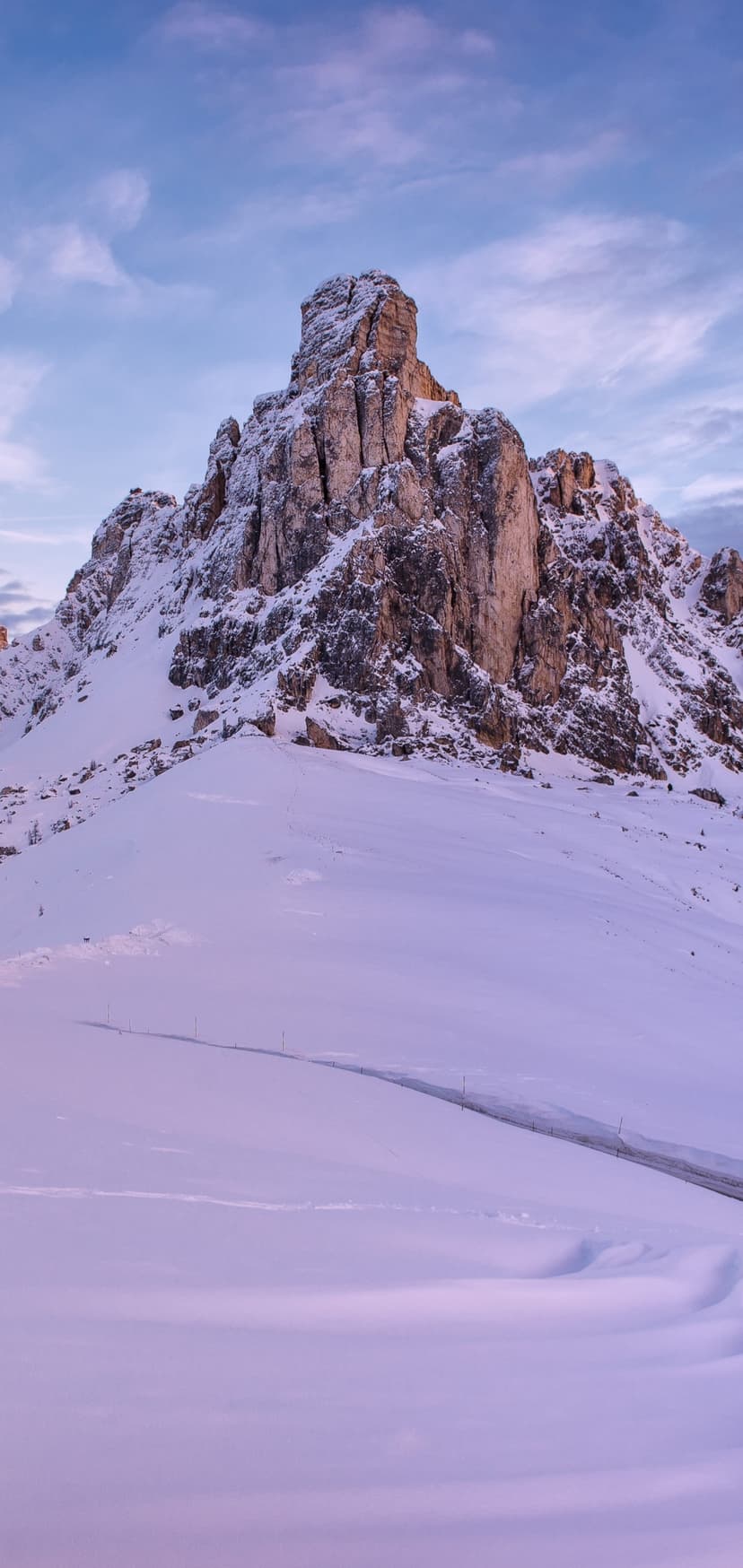 Snow-covered mountain peak towers above a winding alpine road in the Italian Dolomites under a soft purple sky.