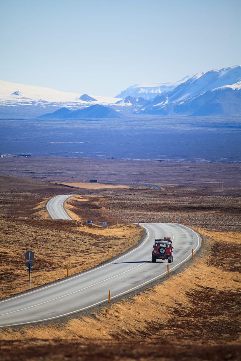 Red off-road vehicle driving on a curved asphalt road toward distant snow-capped mountains under a clear sky.