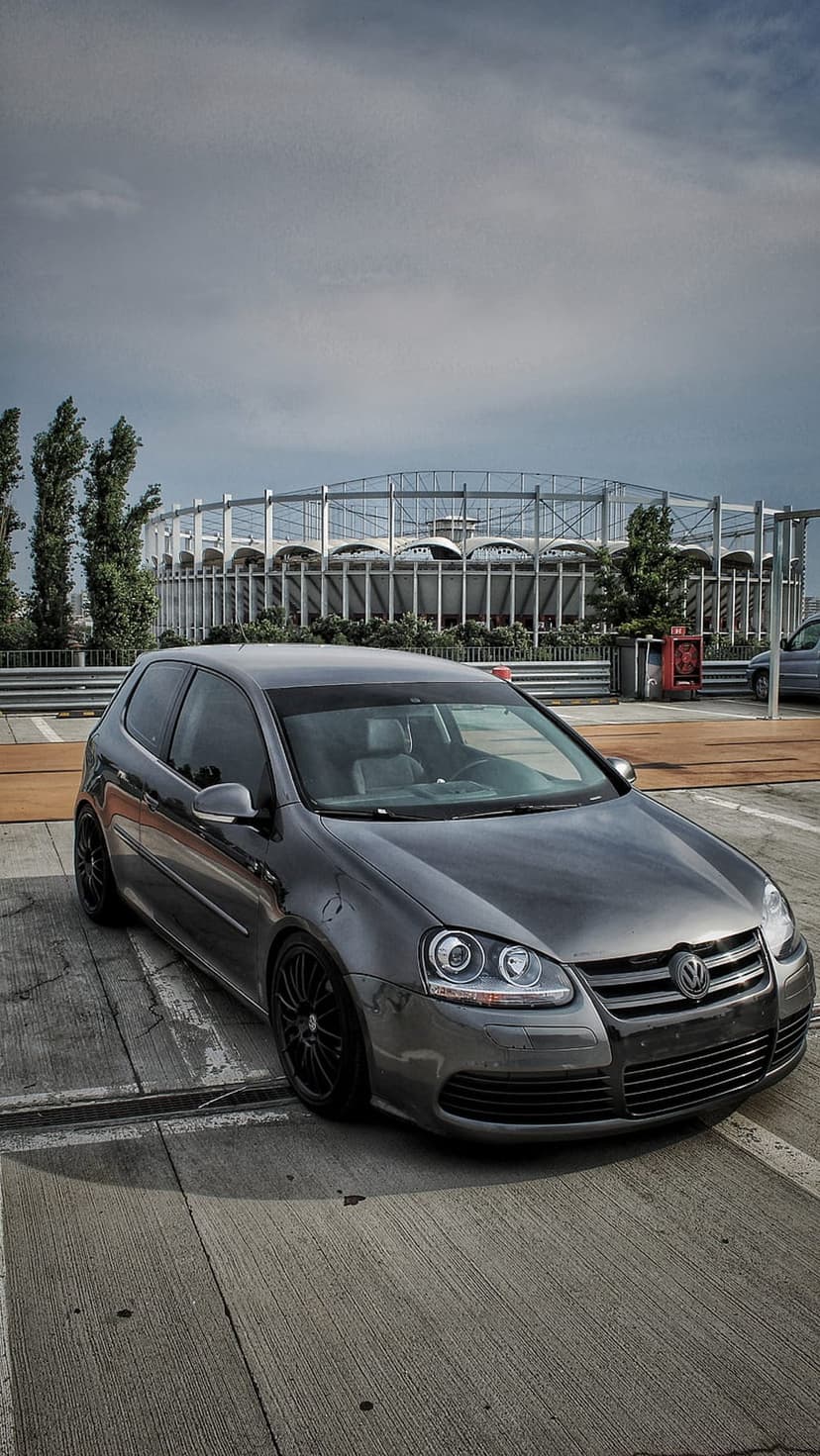 Grey Volkswagen Golf GTI with black wheels parked on concrete in front of a white metal stadium under a cloudy sky.