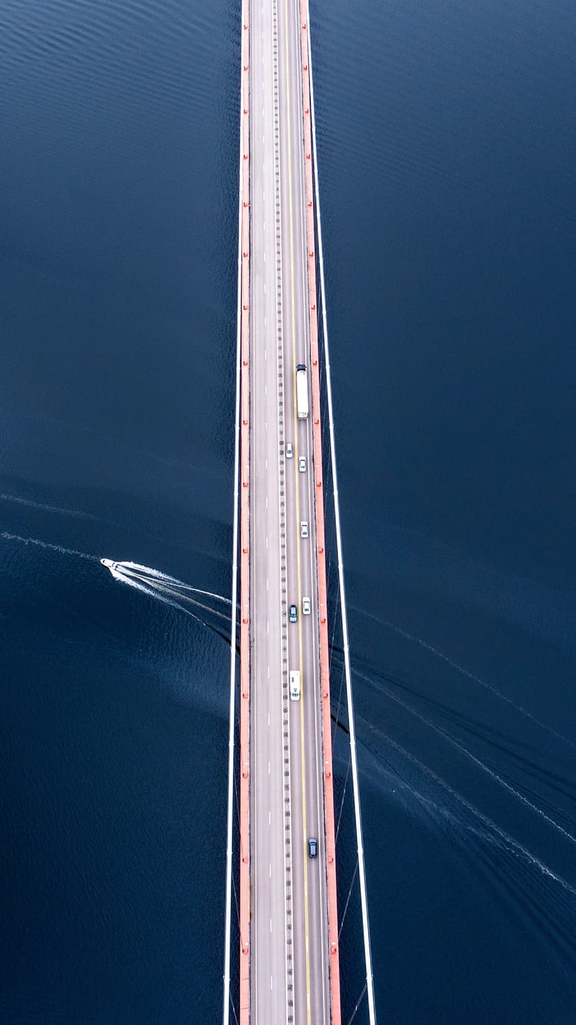 Modern bridge with red supports crosses deep blue water with cars and a white speedboat wake seen from above.