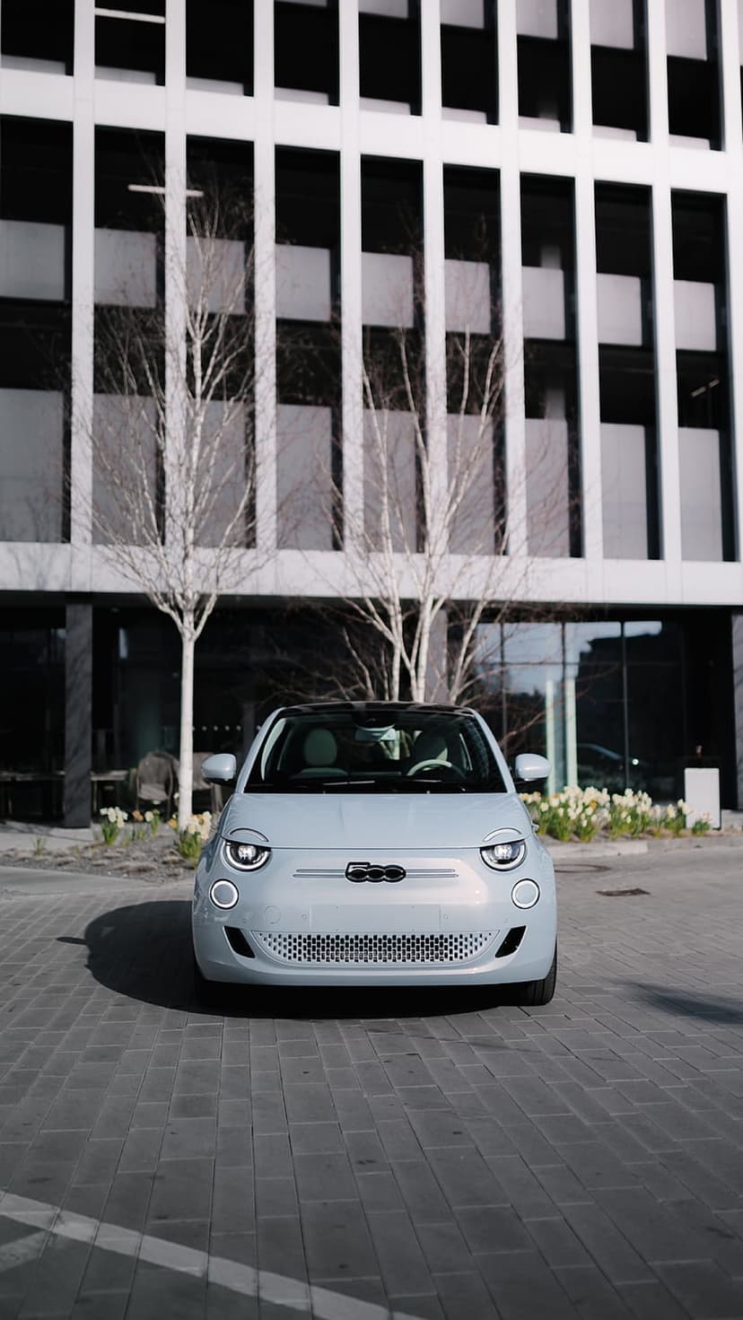 Pale blue Fiat 500 electric car with round headlights parked on bricks near a modern glass building.