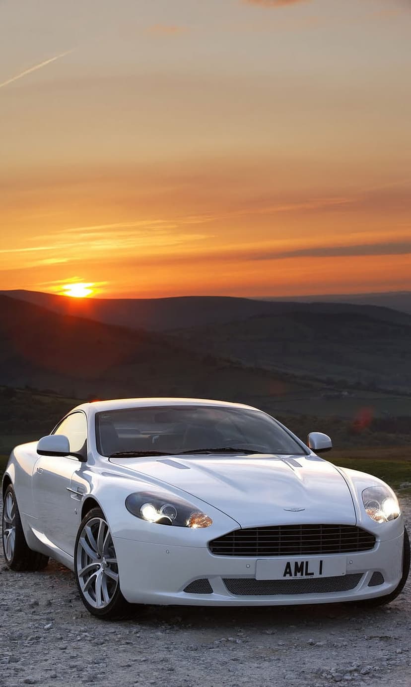 White Aston Martin DB9 parked on a gravel path against a sunset sky with orange clouds and rolling green hills.
