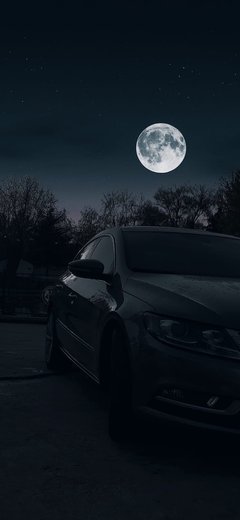 Dark car parked in an open field under a large full moon, twinkling stars, and silhouetted forest trees.