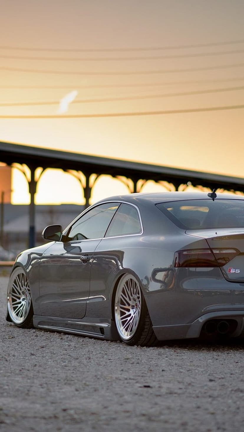 Audi S5 coupe with custom wheels and lowered suspension parked at sunset against an industrial backdrop.