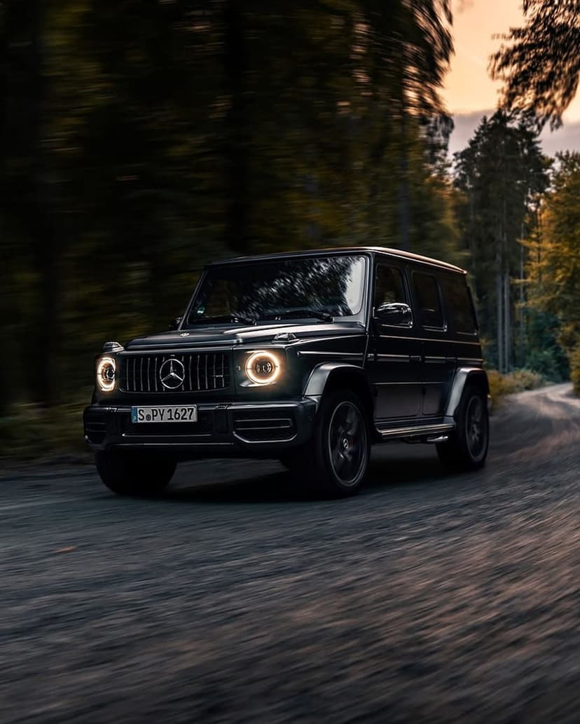 Black Mercedes-Benz G-Wagon driving on a winding forest road with glowing headlights and lush green trees.