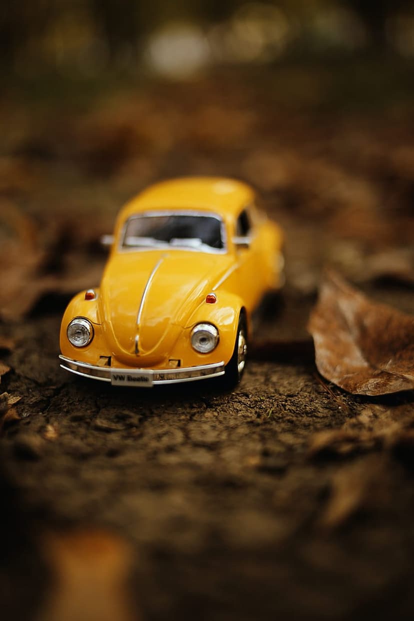 Yellow Beetle toy car parked on brown autumn leaves with a soft-focus background in a vertical phone orientation.
