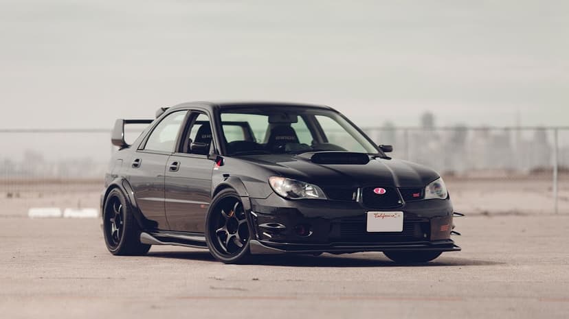 Modified black Subaru WRX STI with widebody kit and racing seats parked on asphalt against a blurred city skyline.