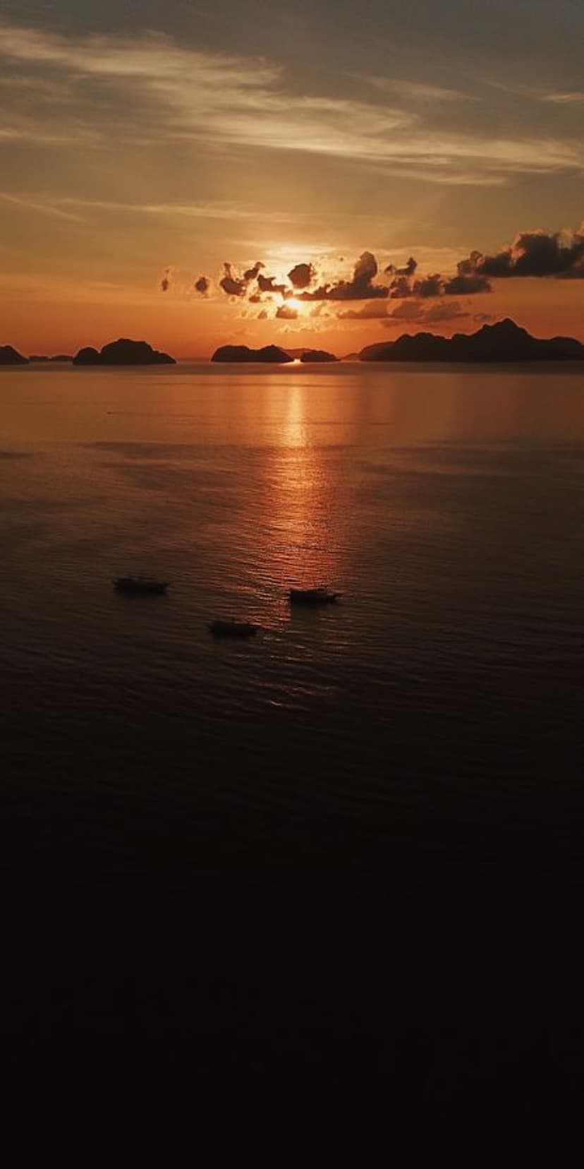 Sun setting over tropical waters with silhouetted boats and islands reflecting a vibrant golden orange sky.