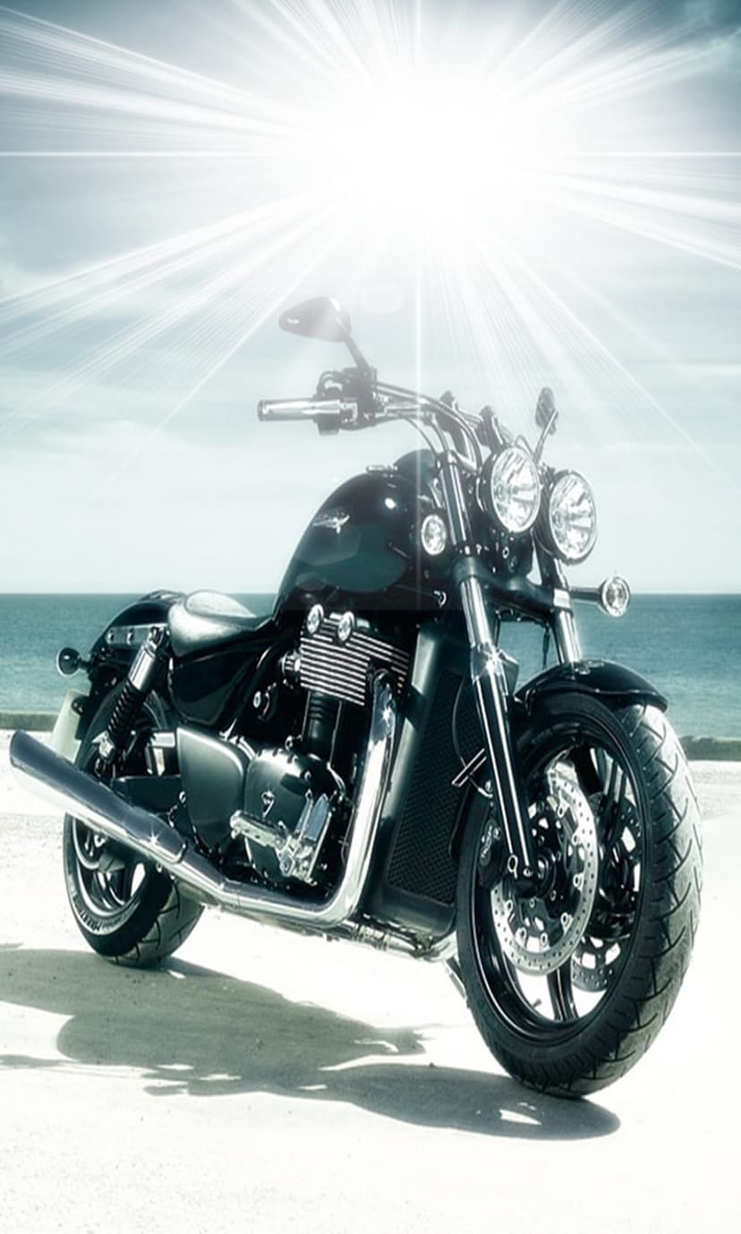 Black Triumph motorcycle parked on a sandy beach with turquoise ocean waves and a bright sun flare in the sky.