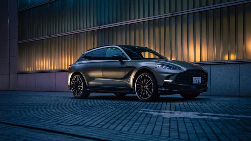 Matte bronze luxury SUV parked on a paved street in front of a glowing architectural glass building at night.