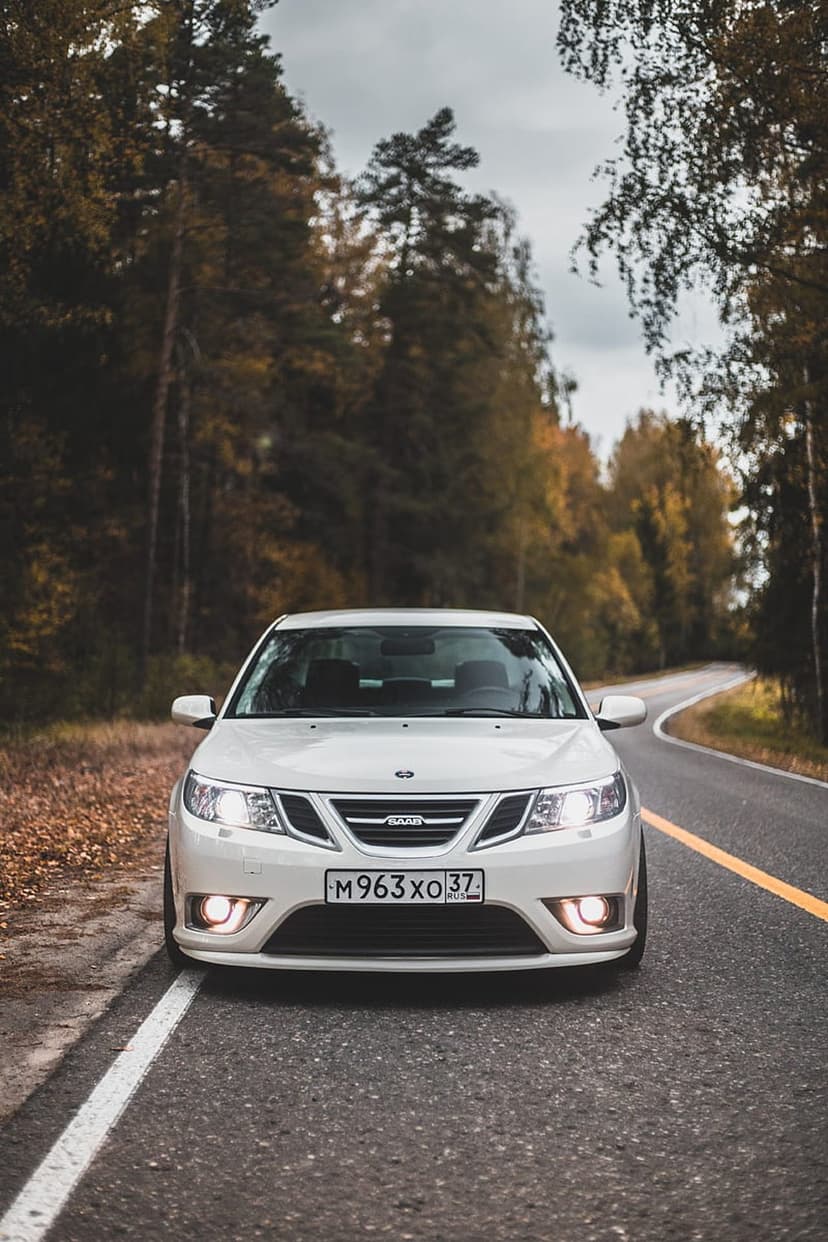 White Saab 9-3 sedan with glowing headlights drives on a winding road through dense autumn trees and orange leaves.