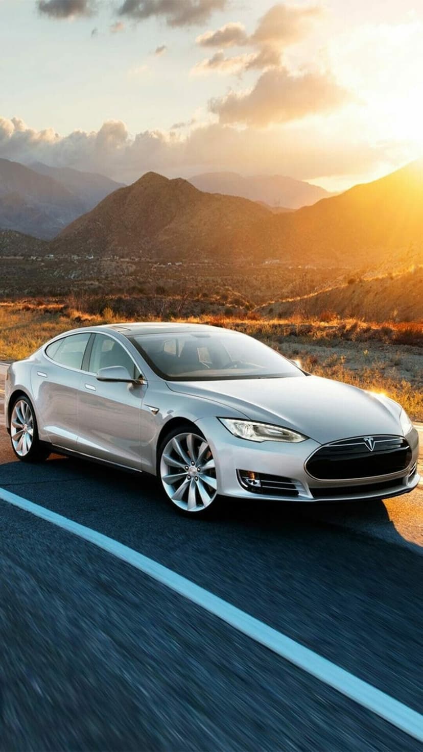 Silver Tesla Model S driving on a mountain road at sunset with golden lighting and a scenic landscape background.