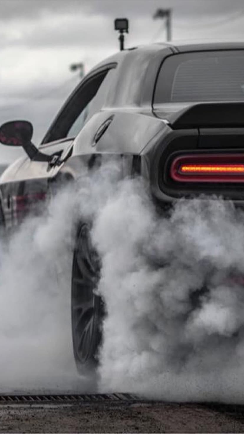 Black muscle car emits thick white tire smoke during a burnout with a glowing red taillight visible in the haze.