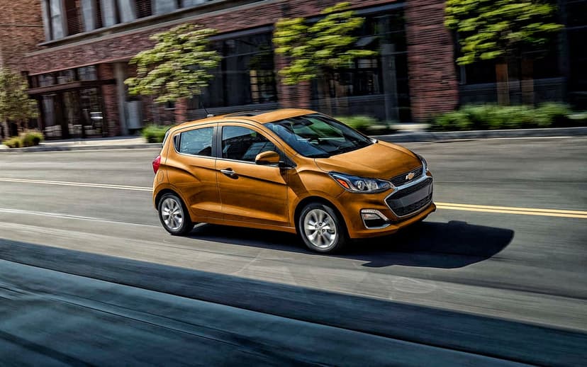 Bronze Chevrolet Spark driving on a city street with motion-blurred brick buildings and trees background.