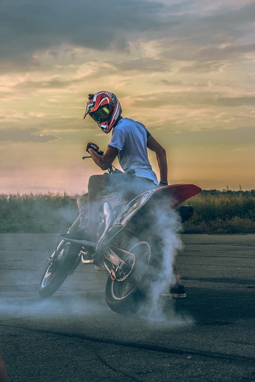 Motorcyclist on a dirt bike performs a burnout with thick white smoke against a dramatic orange sunset sky.