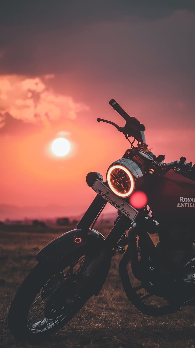Royal Enfield motorcycle silhouette with a glowing round headlight against an orange and pink sunset sky background.