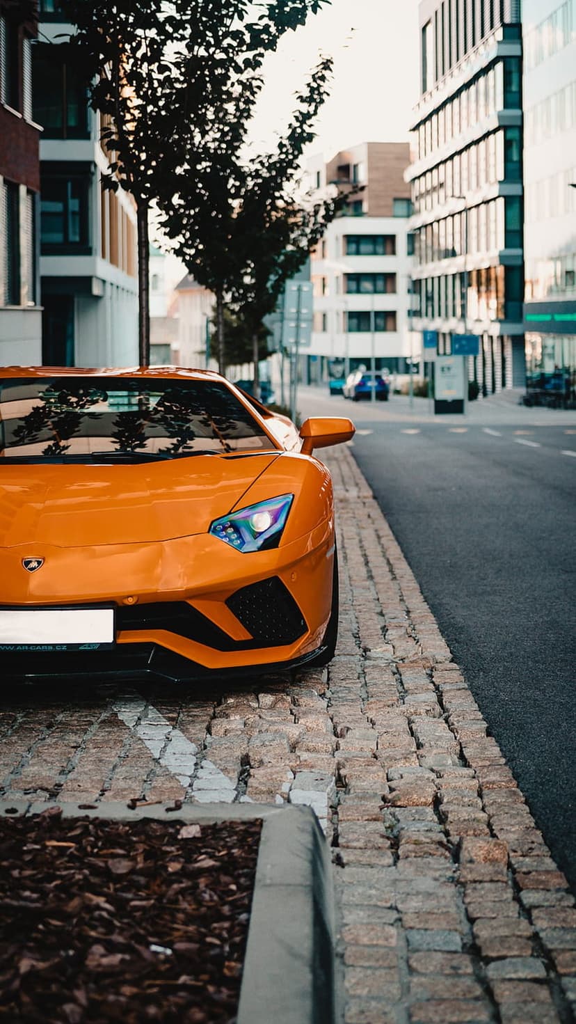 Orange Lamborghini Aventador with sharp headlights parked on a gray cobblestone street in front of modern buildings.