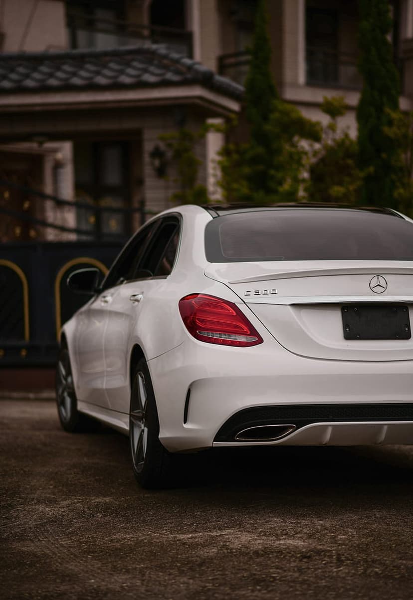 White Mercedes-Benz C-Class sedan with a black sunroof parked on a gravel drive before an ornate luxury mansion.