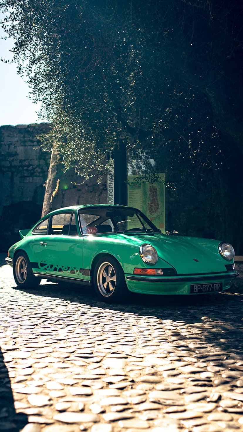 Emerald green Porsche Carrera parked on a sun-dappled cobblestone street with shadows from overhead tree leaves.