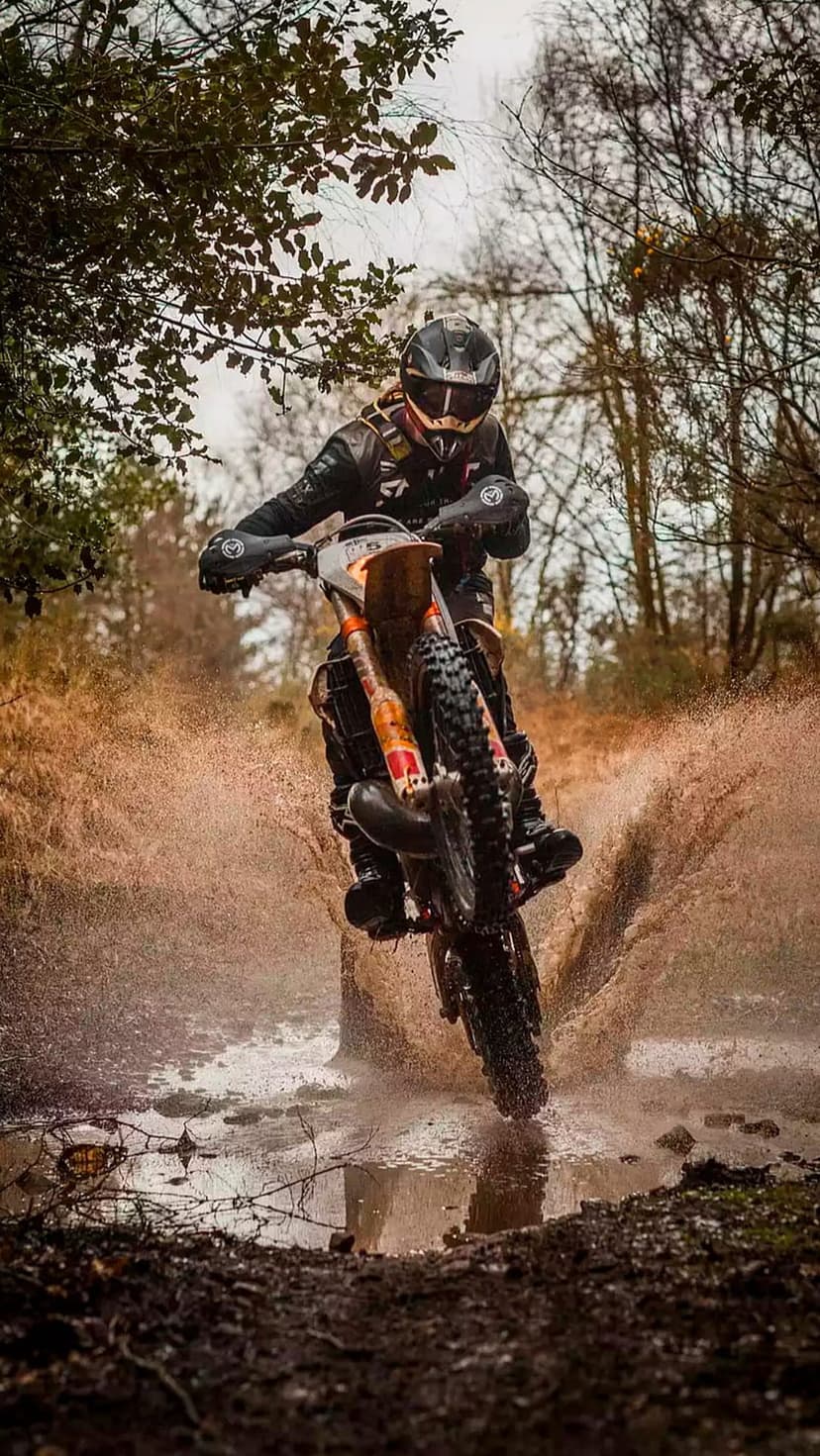 Motocross rider in gear jumps through a deep puddle creating a dramatic spray of mud and water in a forest.