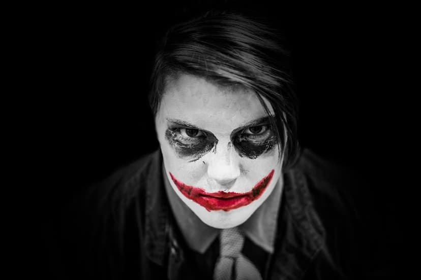 Joker-style face paint with a red smile and white skin against a black background in a moody close-up portrait.