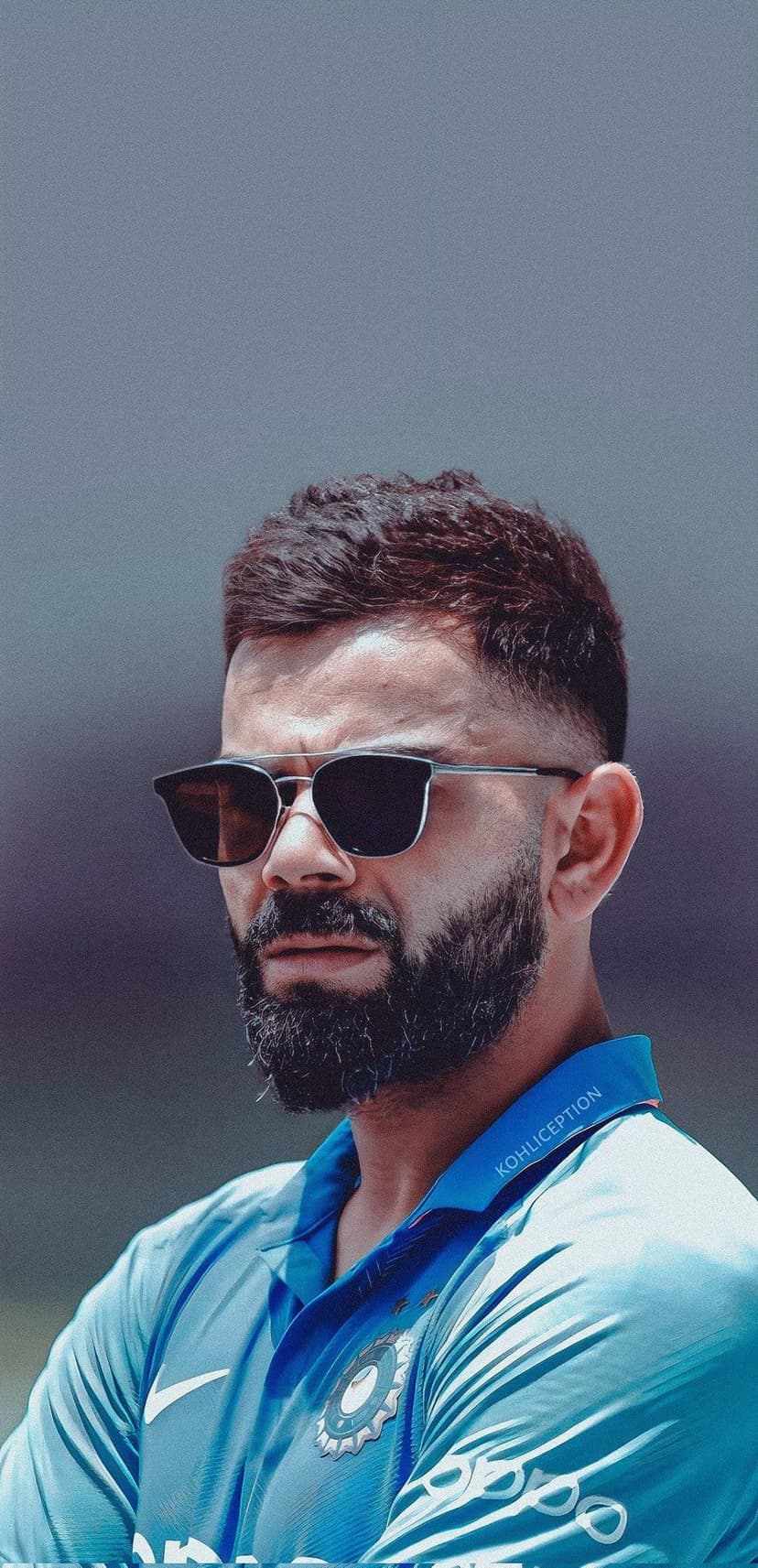 Virat Kohli wearing a blue cricket jersey and dark sunglasses with a visible beard in a close-up shot.
