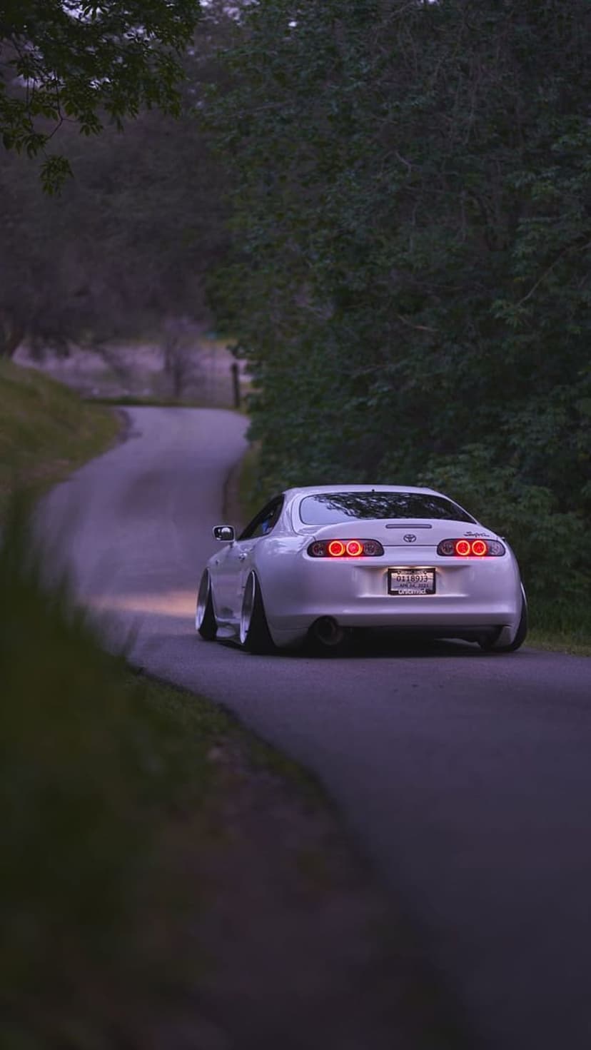White Toyota Supra with glowing red taillights driving on a winding forest road during a misty twilight evening.