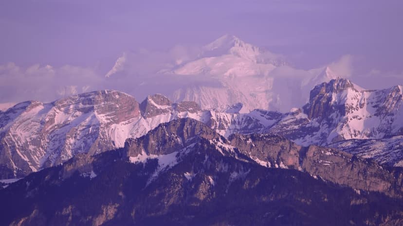 Snow-capped mountain peaks emerge from clouds beneath a violet sky during a tranquil alpine dusk or dawn.