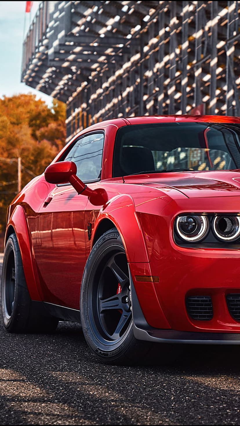 Red Dodge Challenger Hellcat Widebody parked on asphalt near a lattice building and autumn leaves.