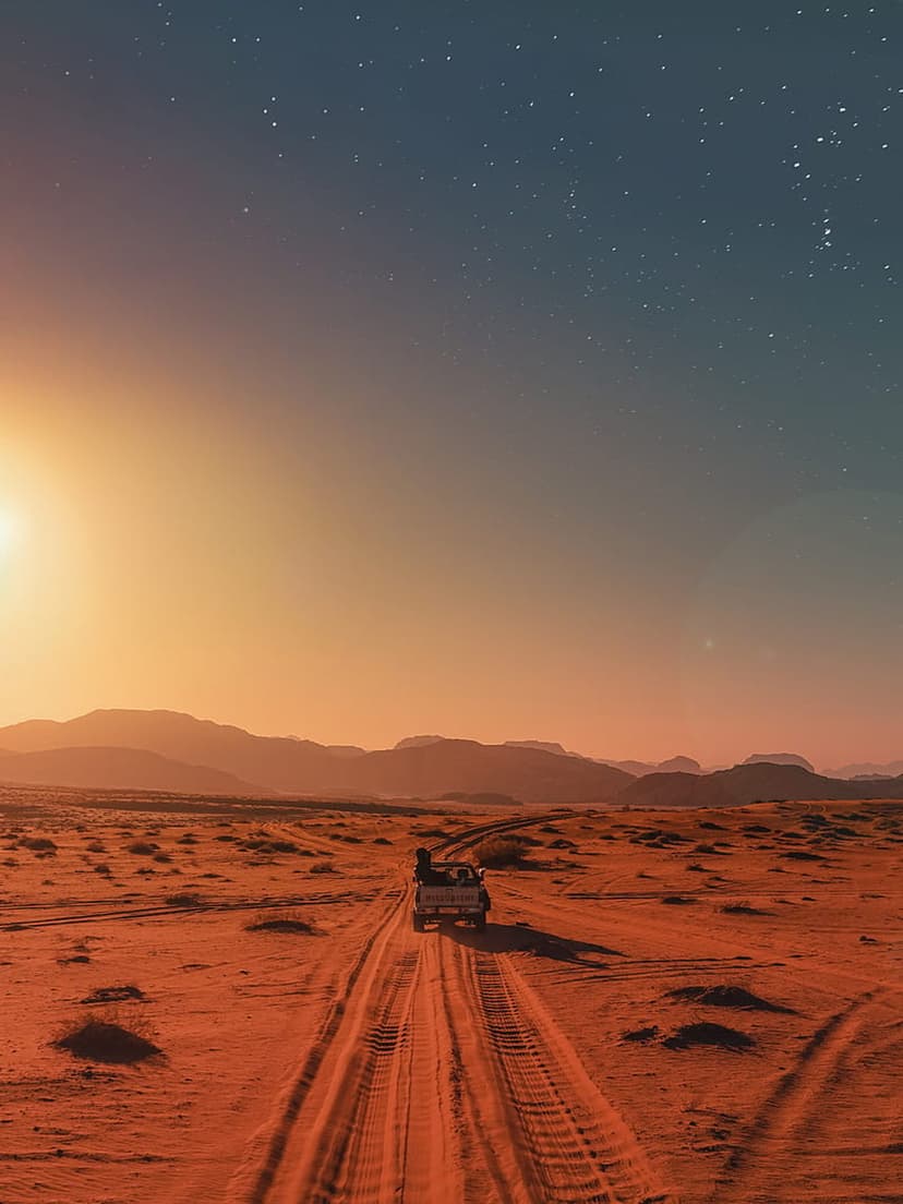 Off-road jeep traveling across orange sand dunes at sunset beneath a clear sky filled with bright stars.