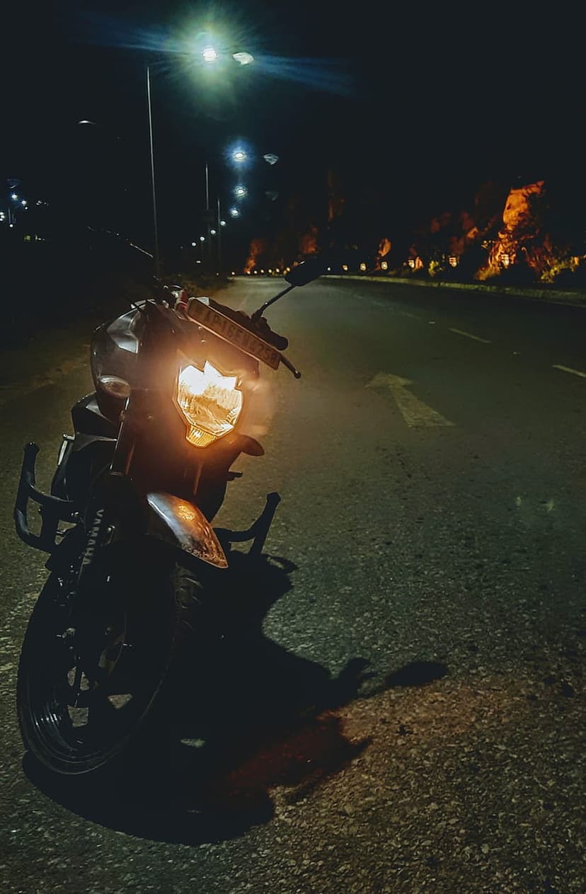 Motorcycle headlight illuminates a dark asphalt highway lined with streetlights and silhouettes of distant trees.