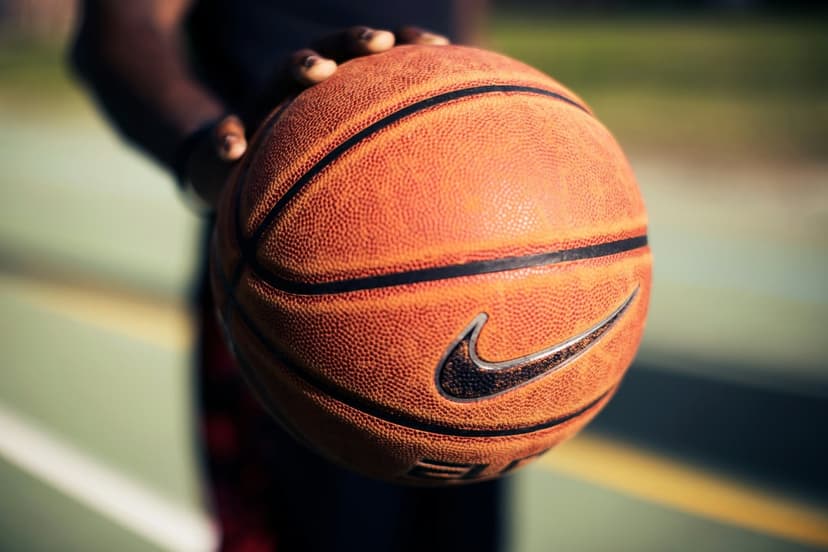 Hand holding a textured Nike basketball with a visible swoosh logo on a blurred outdoor court during the daytime.