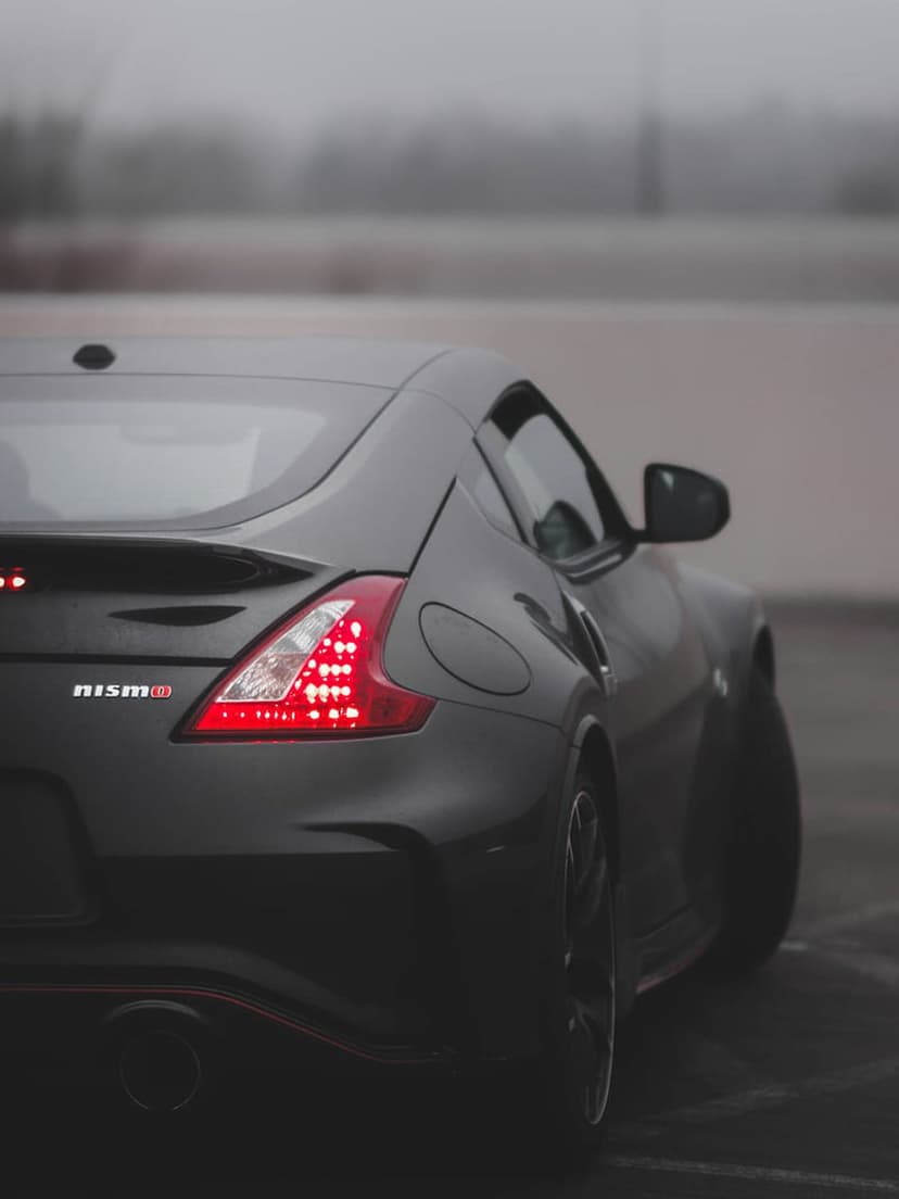 Nissan 370Z Nismo rear taillight with glowing red LEDs on a black car body parked on a misty asphalt road surface.