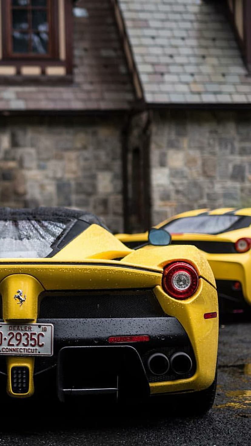 Two yellow Ferrari sports cars parked on wet asphalt in front of a stone building with slate roof tiles.