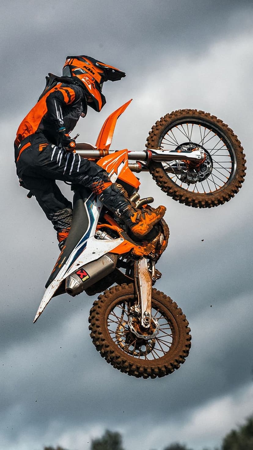 Motocross rider in orange gear jumps a dirt bike against a gray cloudy sky with mud visible on the tires.