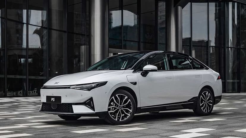 White Xpeng P5 electric sedan on a checkered stone plaza in front of a modern glass building with deep reflections.