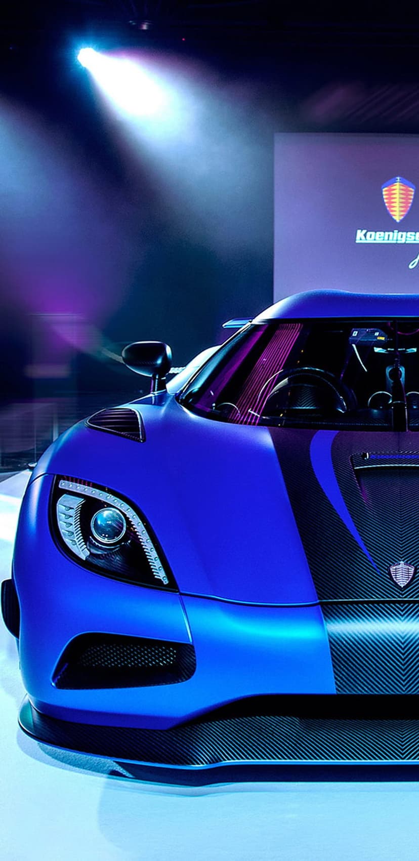 Blue Koenigsegg hypercar with glowing headlights stands on a stage before a purple logo wall in vertical view.