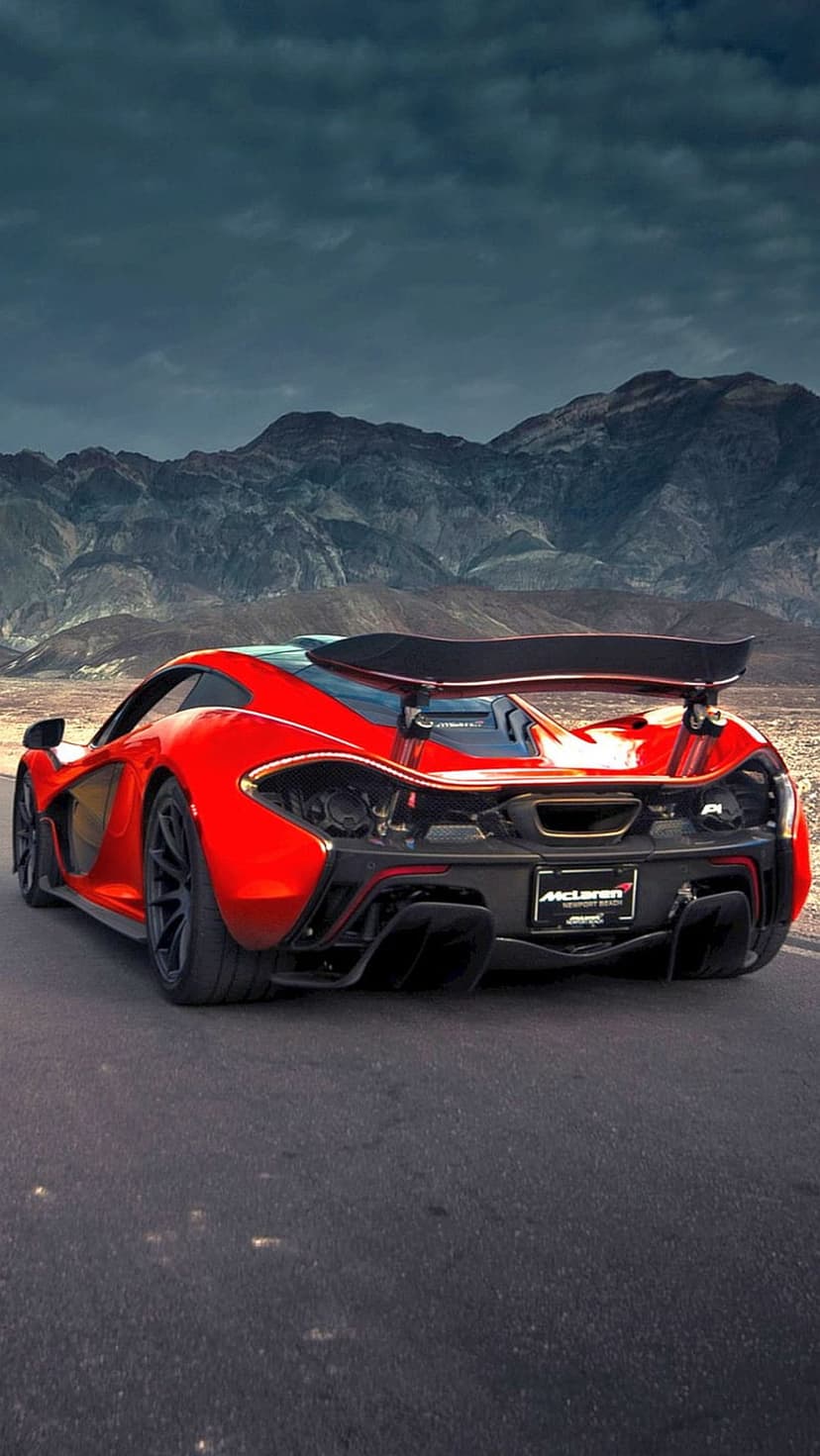 Red McLaren P1 supercar drives on a desert road against a backdrop of rugged mountains and dark clouds.