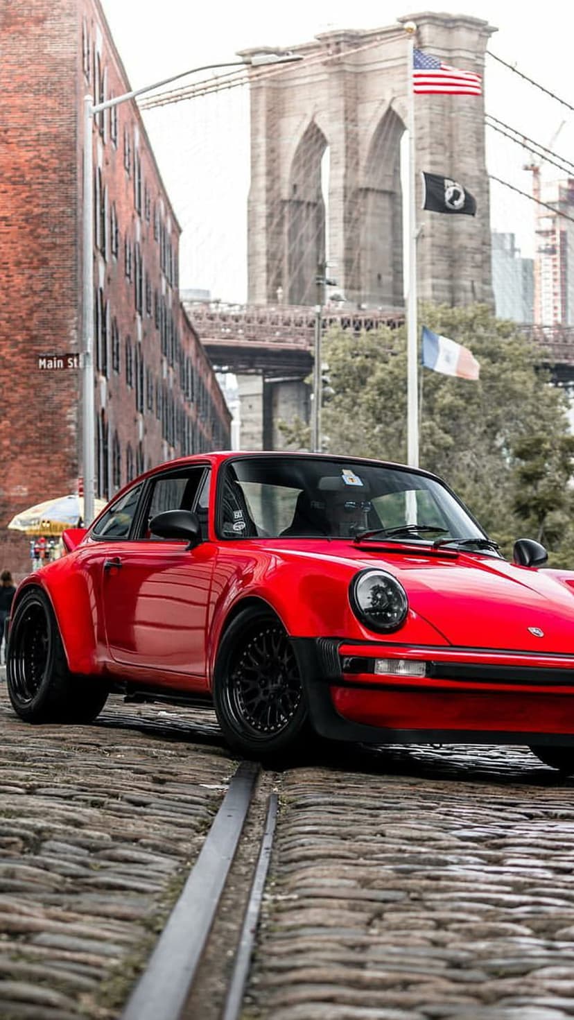 Red Porsche 911 Turbo parked on a cobblestone street with the Brooklyn Bridge and US flags in the background.
