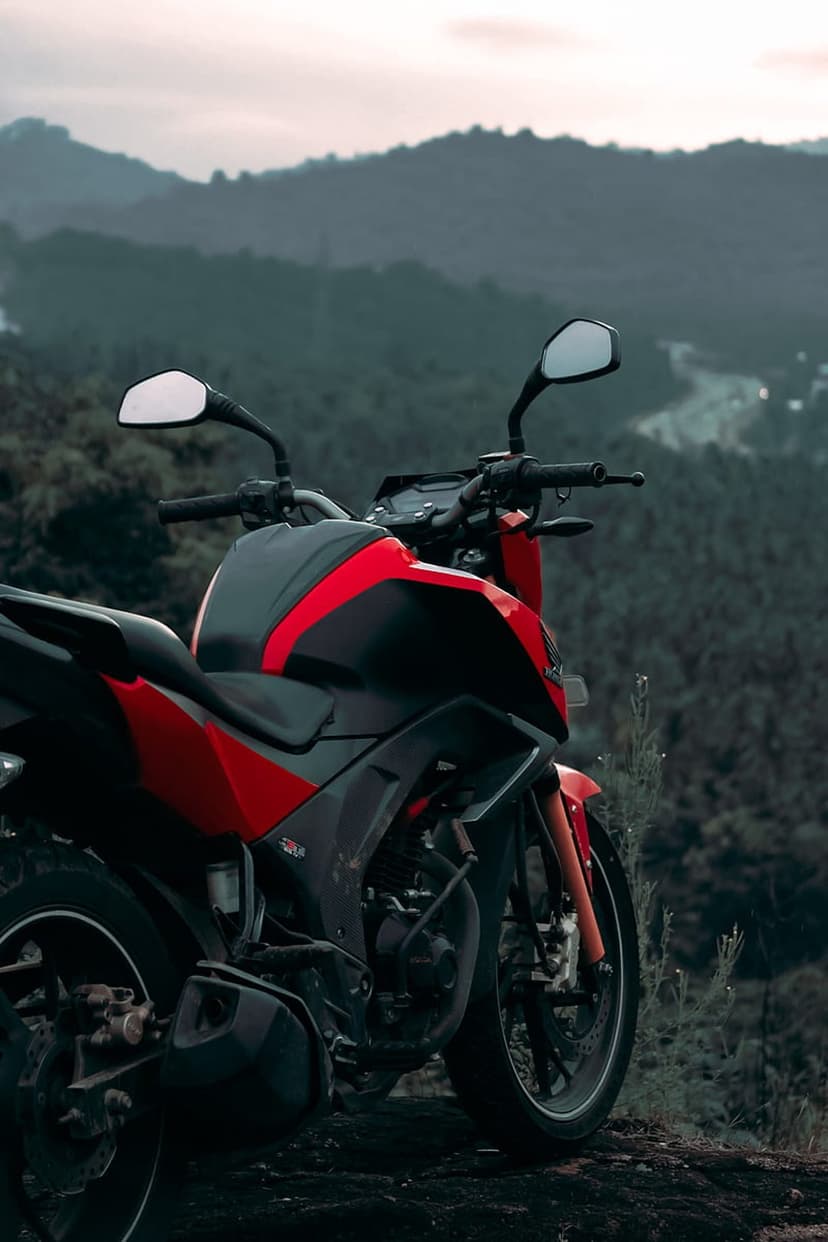 Red and black sportbike parked on a rocky cliff edge overlooking misty green mountain peaks under an overcast sky.