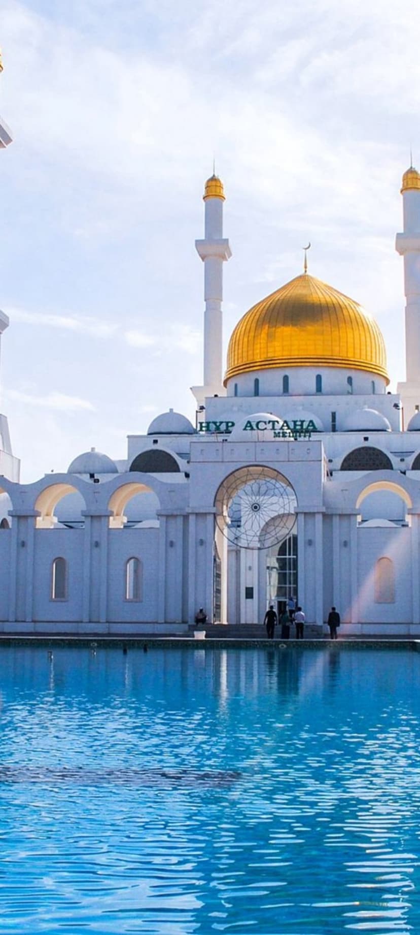 Nur Astana Mosque with a gold dome and white minarets reflects in a blue pool under a bright sunny sky.