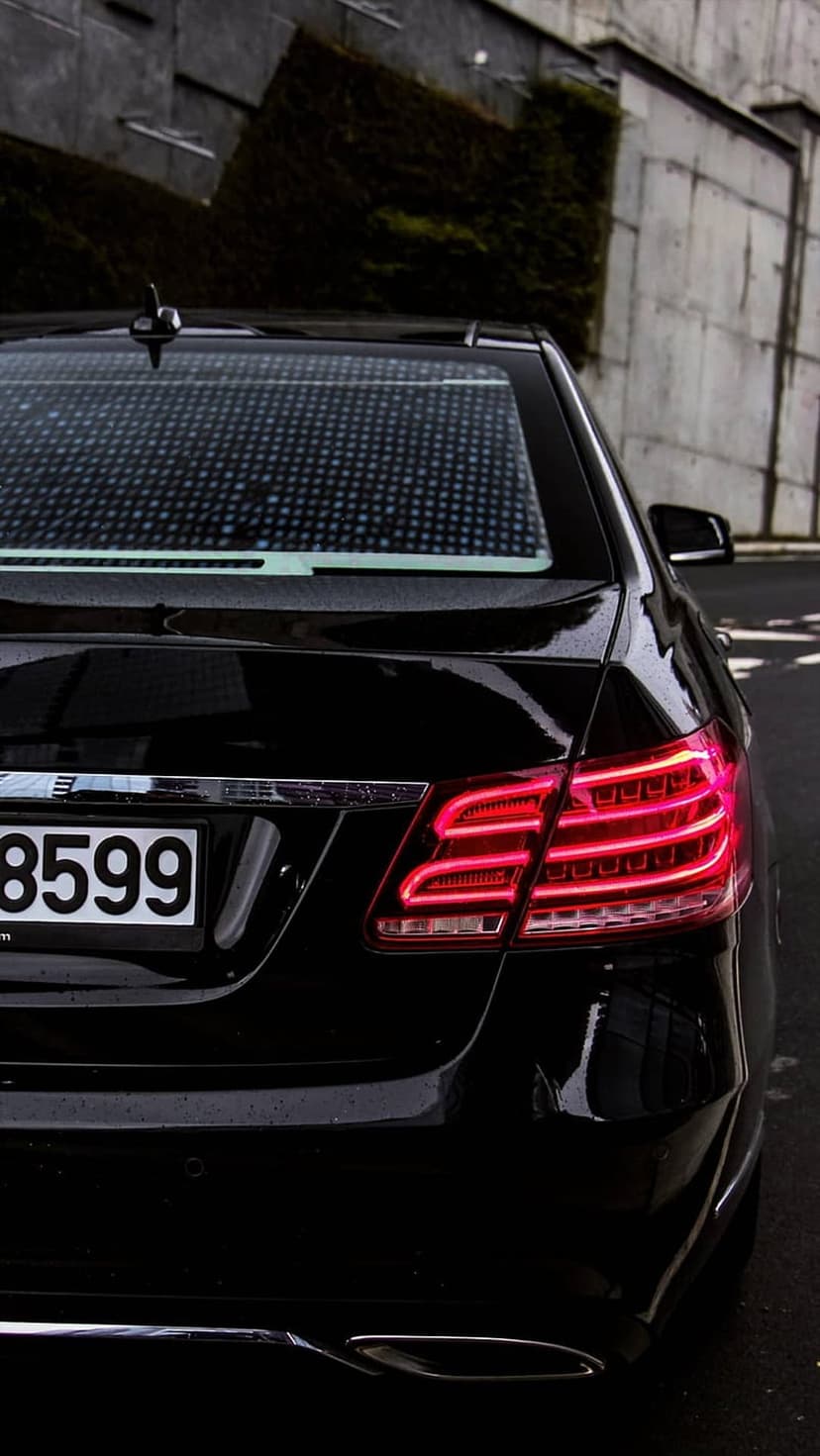 Mercedes E-Class LED tail lights glow red next to a license plate against a textured grey concrete wall backdrop.