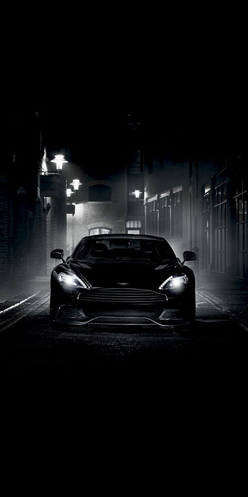 Black Aston Martin sports car parked in a dark rainy alleyway between brick buildings under amber streetlights.