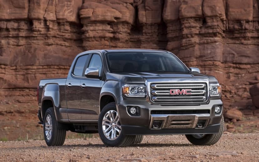 Dark gray GMC Canyon pickup truck parked on desert ground with massive red sandstone cliffs behind it.