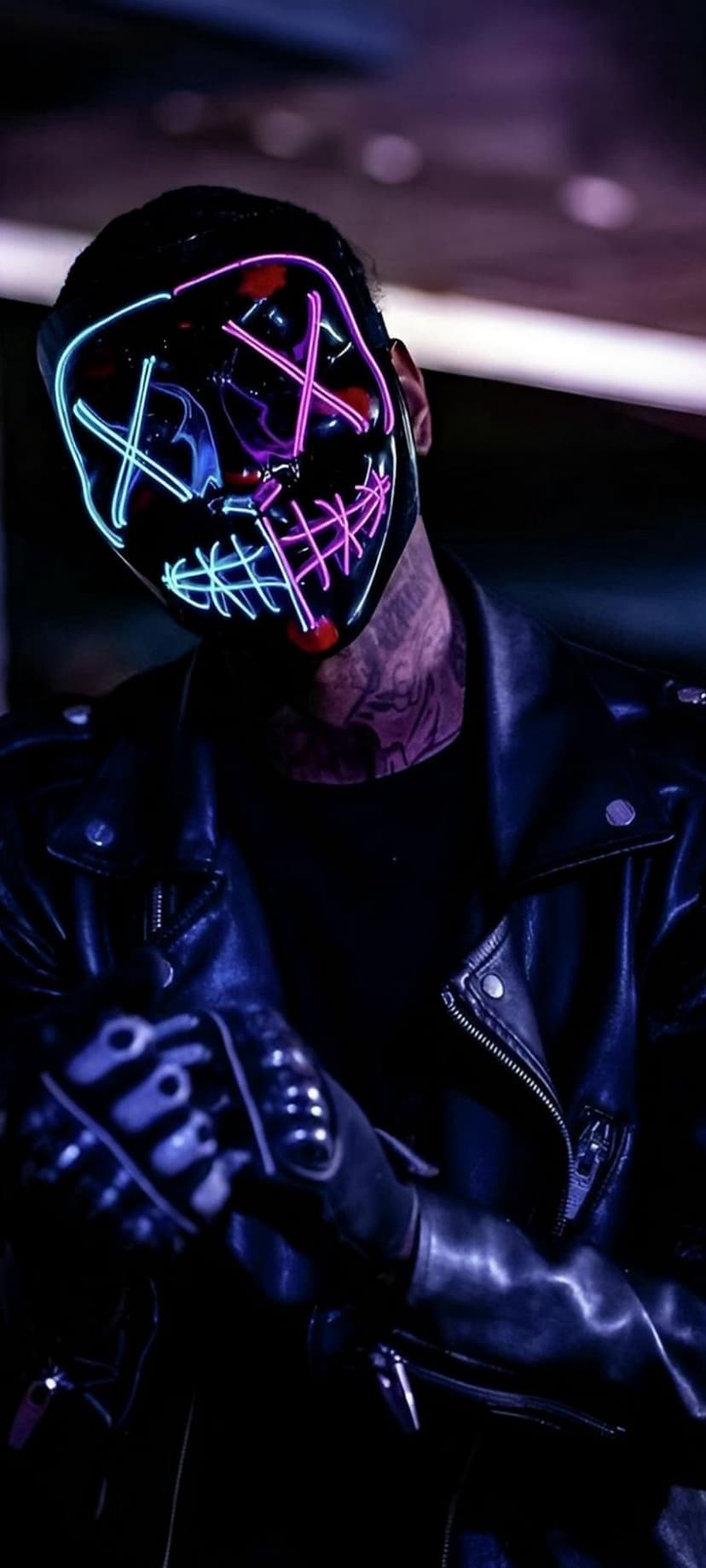 Man wearing a glowing blue neon purge mask and black leather jacket against a dark, moody urban backdrop.