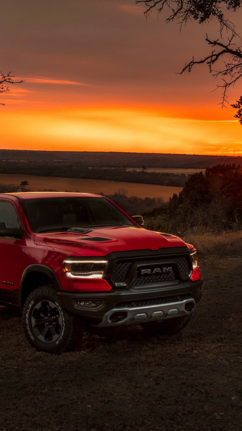 Red Ram 1500 truck with glowing headlights parked on a grassy ridge against a fiery orange sunset sky.