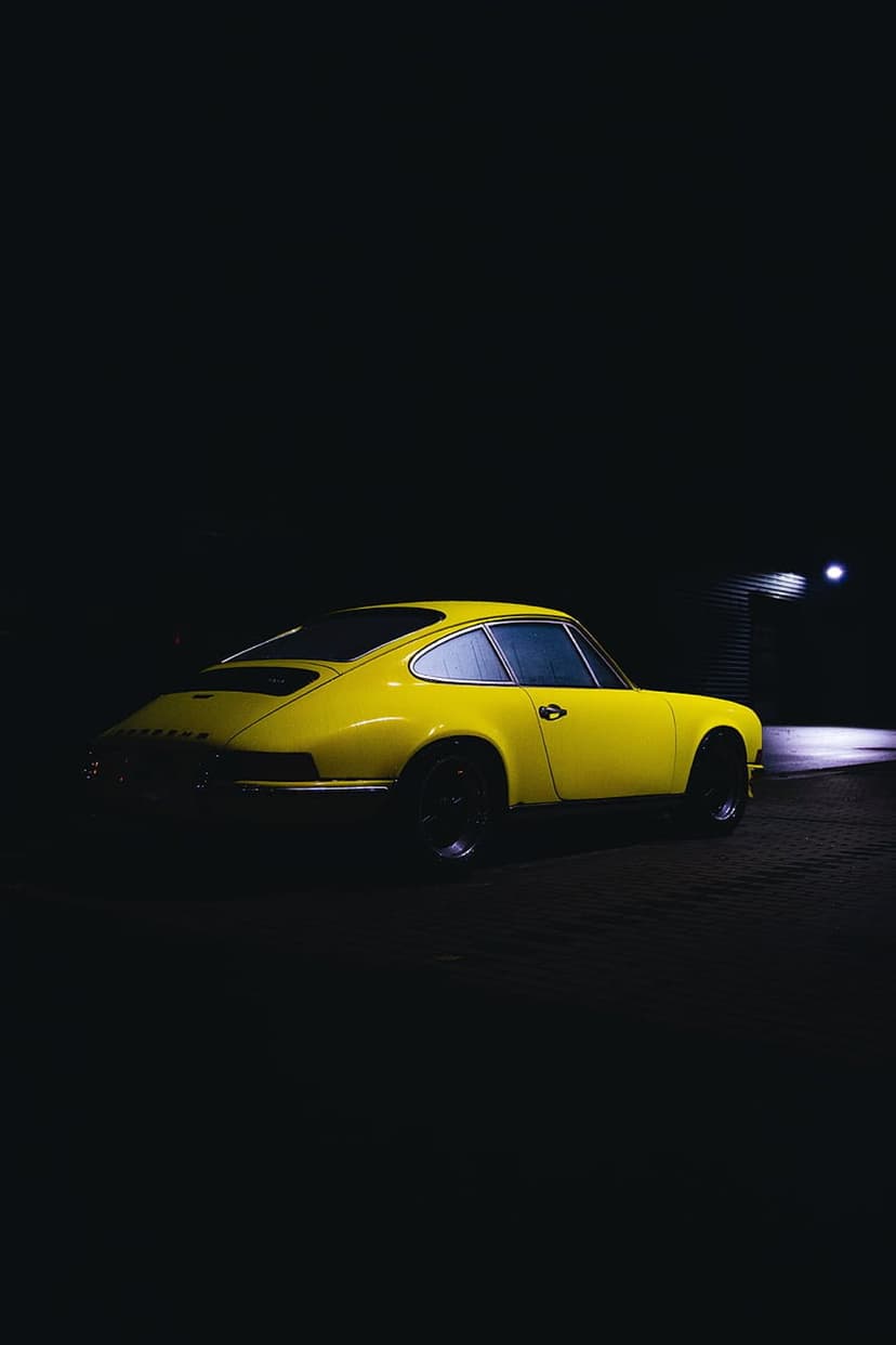 Yellow vintage Porsche 911 coupe parked on dark pavement at night with dramatic side lighting from the side.
