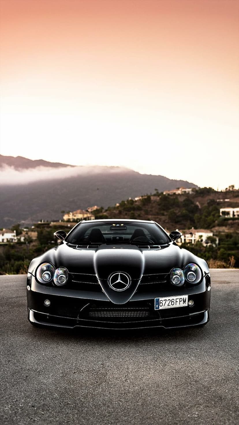 Black Mercedes SLR McLaren Roadster parked on a road with fog-covered hills and a sunset in the background.