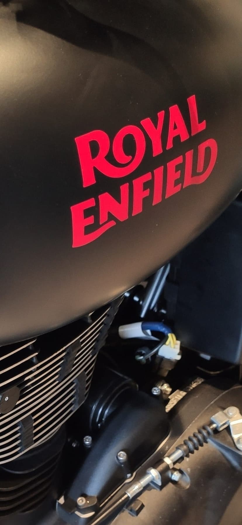 Matte black motorcycle fuel tank featuring a red Royal Enfield logo above a detailed engine and wiring.