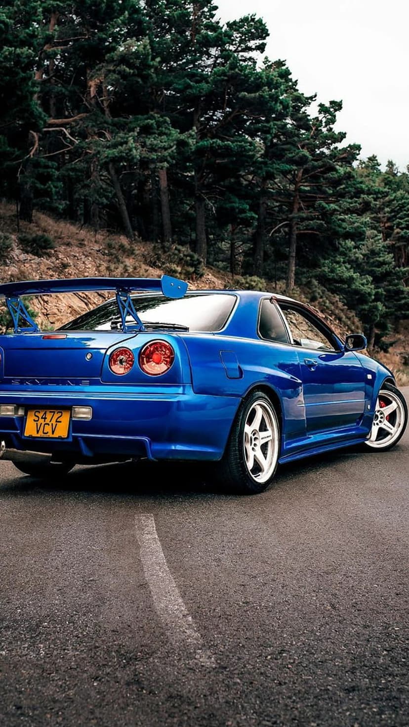Blue Nissan Skyline R34 GT-R with quad taillights parked on a paved road surrounded by tall pine trees.